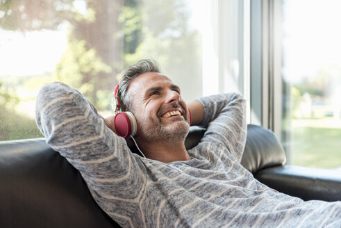 Happy mature man lying on couch at home wearing headphones - DIGF04389