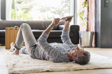 Mature man lying on carpet at home using a tablet - DIGF04390