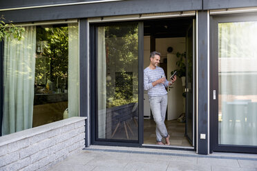 Smiling mature standing at French door at home using a tablet - DIGF04395