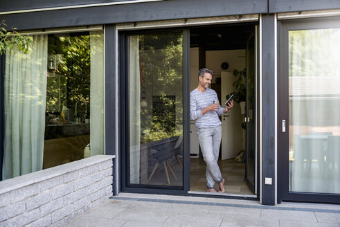 Lächelnde Reife, die zu Hause an der Terrassentür steht und ein Tablet benutzt - DIGF04395