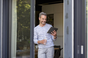 Lächelnde Reife, die zu Hause an der Terrassentür steht und ein Tablet benutzt - DIGF04396