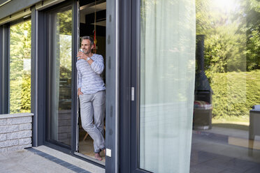 Smiling mature standing at French door at home drinking from glass - DIGF04397