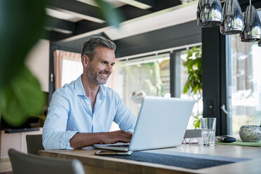 Smiling mature man at home using a laptop at table - DIGF04419