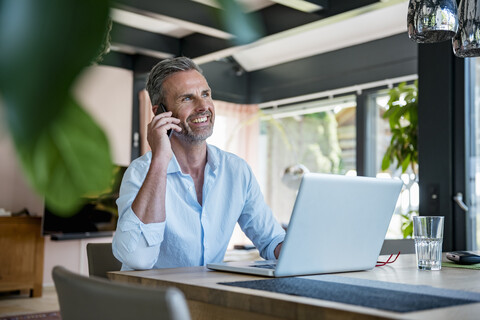 Lächelnder reifer Mann zu Hause mit Handy und Laptop am Tisch, lizenzfreies Stockfoto