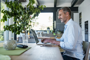 Smiling mature man at home using a laptop at table - DIGF04421