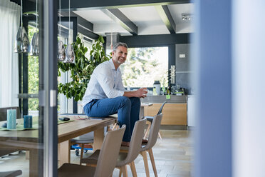 Smiling mature man at home sitting on table with cup of coffee - DIGF04422