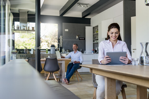 Smiling woman at home using a tablet at table with man in background using laptop stock photo