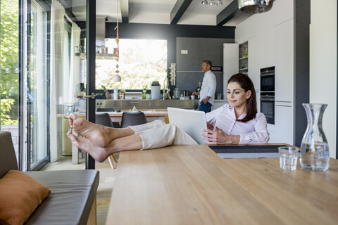 Entspannte Frau zu Hause mit einem Laptop am Tisch mit einem Mann im Hintergrund, lizenzfreies Stockfoto