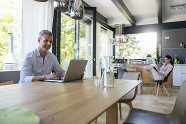 Lächelnder Mann zu Hause, der einen Laptop am Tisch benutzt, mit einer Frau im Hintergrund, die Zeitung liest - DIGF04436