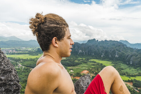 Laos, Vang Vieng, junger Mann auf einem Felsen mit Blick auf eine Landschaft mit Reisfeldern - AFVF00517