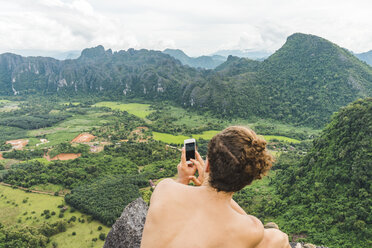 Laos, Vang Vieng, young man on top of rocks taking cell phone picture of landscape - AFVF00518