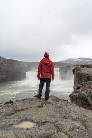 Island, Nordisland, junger Mann schaut zum Wasserfall, lizenzfreies Stockfoto