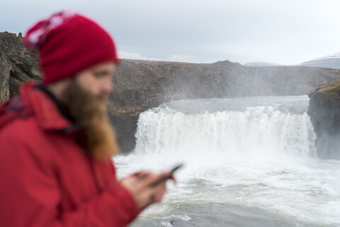 Island, Nordisland, junger Mann mit Smartphone, Wasserfall im Hintergrund, lizenzfreies Stockfoto