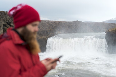 Island, Nordisland, junger Mann mit Smartphone, Wasserfall im Hintergrund - AFVF00535