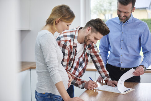 Couple with real estate agent signing contract in kitchen of new apartment - ABIF00452