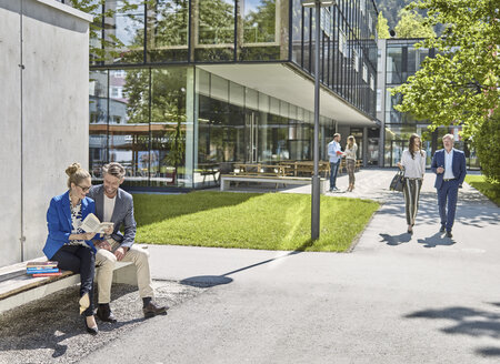 Lächelnde Kollegen mit Buch auf einer Bank vor einem Bürogebäude sitzend - CVF00588