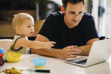 Father and young son sitting at table, father using laptop, son pointing at screen - ISF02366