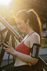 Young female runner wearing earphones and looking at smartphone in sunlight - ISF02439