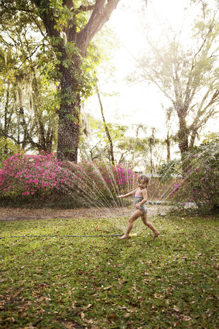 Mädchen im Badeanzug spielt im Wasser des Gartensprinklers, lizenzfreies Stockfoto