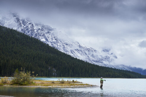 Mann beim Fliegenfischen in einem See neben schneebedeckten Bergen, Banff, Alberta, Kanada - ISF02507