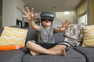 Young boy sitting cross legged on sofa, wearing virtual reality headset, hands reaching out in front of him - ISF02519