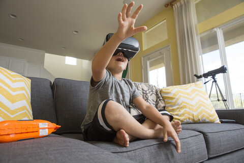 Junge sitzt im Schneidersitz auf dem Sofa, trägt ein Virtual-Reality-Headset und streckt die Hände vor sich aus, lizenzfreies Stockfoto