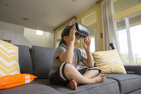 Young boy sitting cross legged on sofa, wearing virtual reality headset - ISF02521