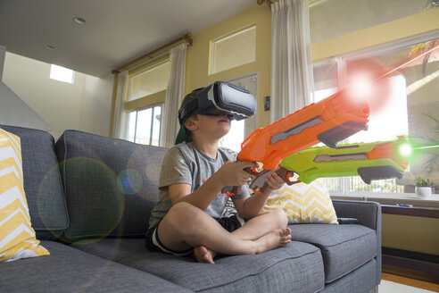 Junge sitzt im Schneidersitz auf dem Sofa, trägt Virtual-Reality-Headset, hält Laserpistolen, digitales Komposit - ISF02522