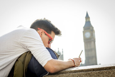 Junger Mann im Freien, an die Wand gelehnt, schaut auf sein Smartphone, Big Ben im Hintergrund, London, England, UK - ISF02595