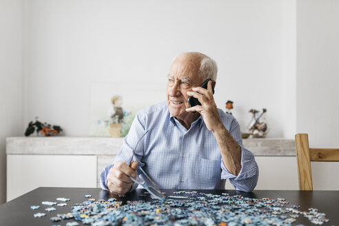 Happy senior man using smartphone while doing a puzzle at home - JRFF01678