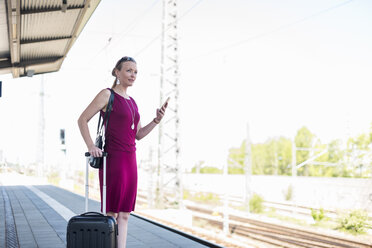 Mature businesswoman with suitcase and smartphone waiting at platform - DIGF04468