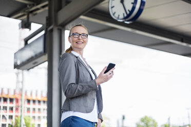 Porträt einer lächelnden Geschäftsfrau mit Mobiltelefon, die am Bahnsteig wartet - DIGF04485