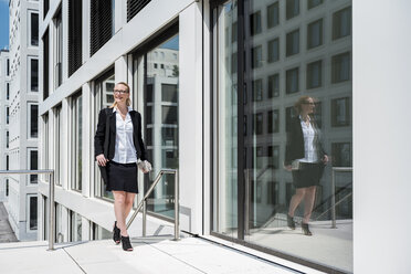 Smiling businesswoman with newspaper walking in front of office building - DIGF04491