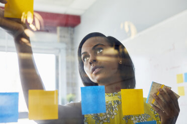 Young woman in office, sticking sticky notes to glass in office - ISF02608