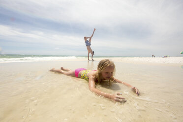 Mädchen liegt am Strand im seichten Wasser, Bruder macht Handstand im Hintergrund - ISF02673