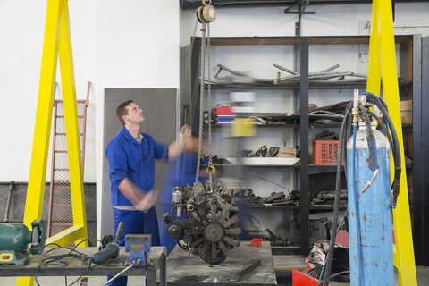 Männlicher Automechaniker, der einen Automotor in einer Reparaturwerkstatt anhebt, lizenzfreies Stockfoto