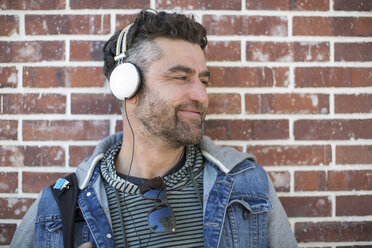 Mid adult man leaning against wall, wearing headphones, looking away, smiling - ISF02964