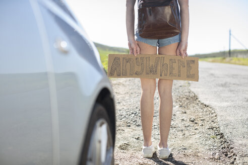 Junge Frau steht neben einem Auto und hält ein Anhalter-Schild mit der Aufschrift 