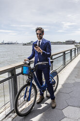 Junger Geschäftsmann auf dem Fahrrad schaut auf sein Smartphone am Ufer des Stadtflusses - ISF03266