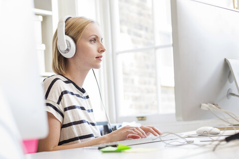 Designerin hört Kopfhörer und tippt am Schreibtisch im Kreativstudio, lizenzfreies Stockfoto