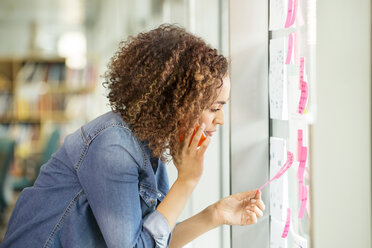 Female digital designer making phone call and looking at adhesive notes on office wall - ISF03385