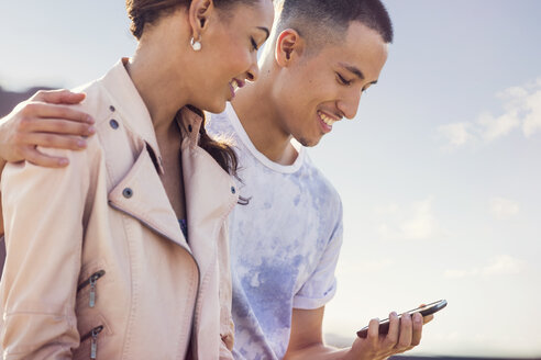 Junges Paar vor blauem Himmel, das bei einer Dachparty auf sein Smartphone schaut - ISF03426
