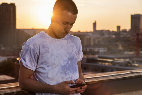 Junger Mann schaut bei Sonnenuntergang auf dem Dach einer Party in London auf sein Smartphone - ISF03435