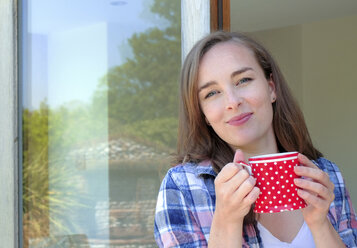 Portrait of young woman holding coffee mug by patio door - ISF03438