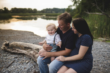 Pregnant couple sitting on beach log with male toddler son, Lake Ontario, Canada - ISF03637