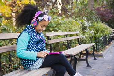 Mädchen sitzt auf einer Parkbank, trägt Kopfhörer und benutzt ein digitales Tablet - ISF03689
