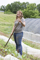 Woman holding hoe in vegetable garden, looking at camera smiling - ISF03749