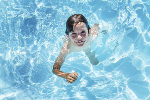 Overhead-Porträt eines Jungen, der in einem von der Sonne beschienenen Schwimmbad im Freien Wasser tritt - ISF03788