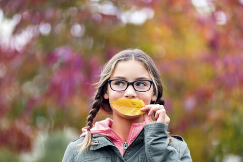 Porträt eines Mädchens mit Zöpfen und Brille, das den Mund mit einem Blatt bedeckt und wegschaut, lizenzfreies Stockfoto