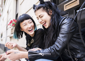Two young stylish female friends sitting doorstep laughing - ISF03819